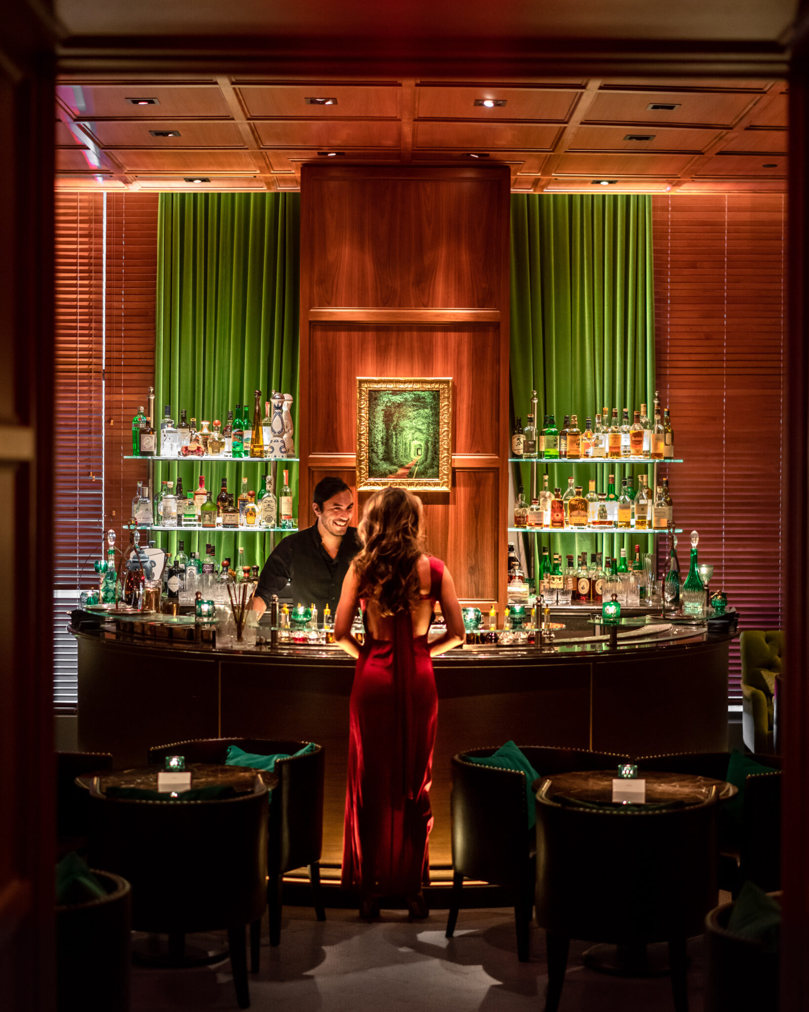 woman in red dress stands at bar talking to bartender in black clothes with green background behind liquor bottles
