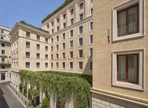 Exterior view of a sand-colored stone building with rows of rectangular windows and vegetation topping the perimeter of a courtyard wall.