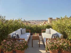 A view of two outdoor couches with wood frames and white upholstery surrounded by potted plants on a roof.
