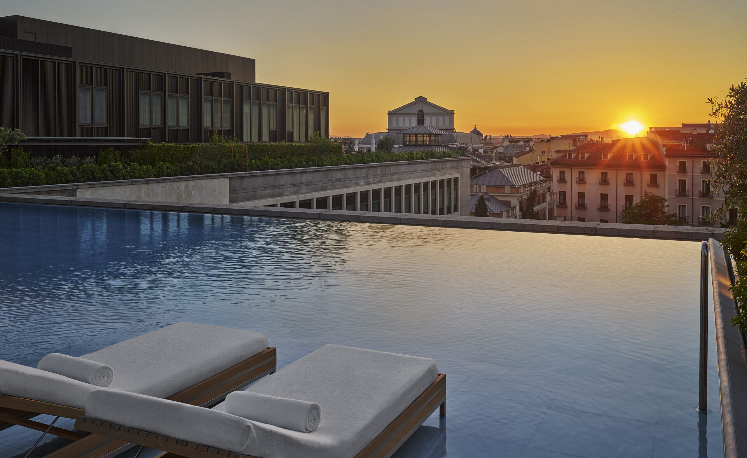 Lounge chairs set inside an outdoor pool with a view of the sun setting over Madrid.